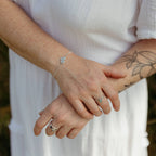 A close-up of a person's hands and arms wearing delicate silver and gemstone stacking rings and a silver mini mantra bracelet with a cluster of flattened silver circles, one with the word 'trust' stamped onto it.  Styled with a white textured dress.