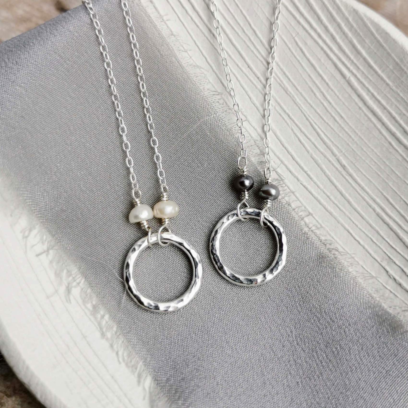 Two hammered silver circle necklaces with pearl details on a gray fabric background
