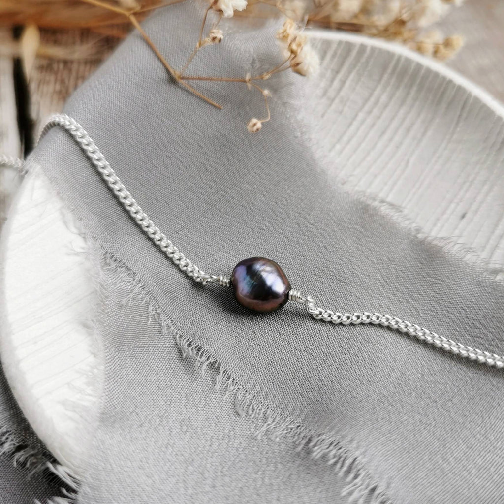 A handmade silver chain bracelet featuring a single freshwater baroque pearl with a dark peacock purple-black body and iridescent highlights, displayed on a textured gray fabric surface.