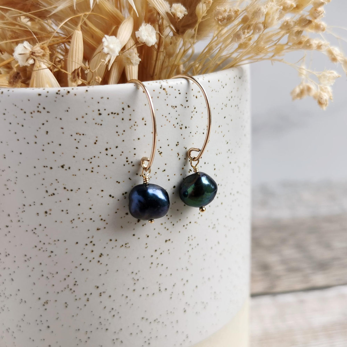 A pair of gold earrings with dark blue-black peacock baroque pearls with iridescent sheen, displayed hanging from the rim of a speckled white ceramic pot containing dried flowers.