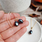 handmade Gold earrings with iridescent black pearls held in a hand, with a blurred background of the matching necklace on a white dish and autumnal decorative items.