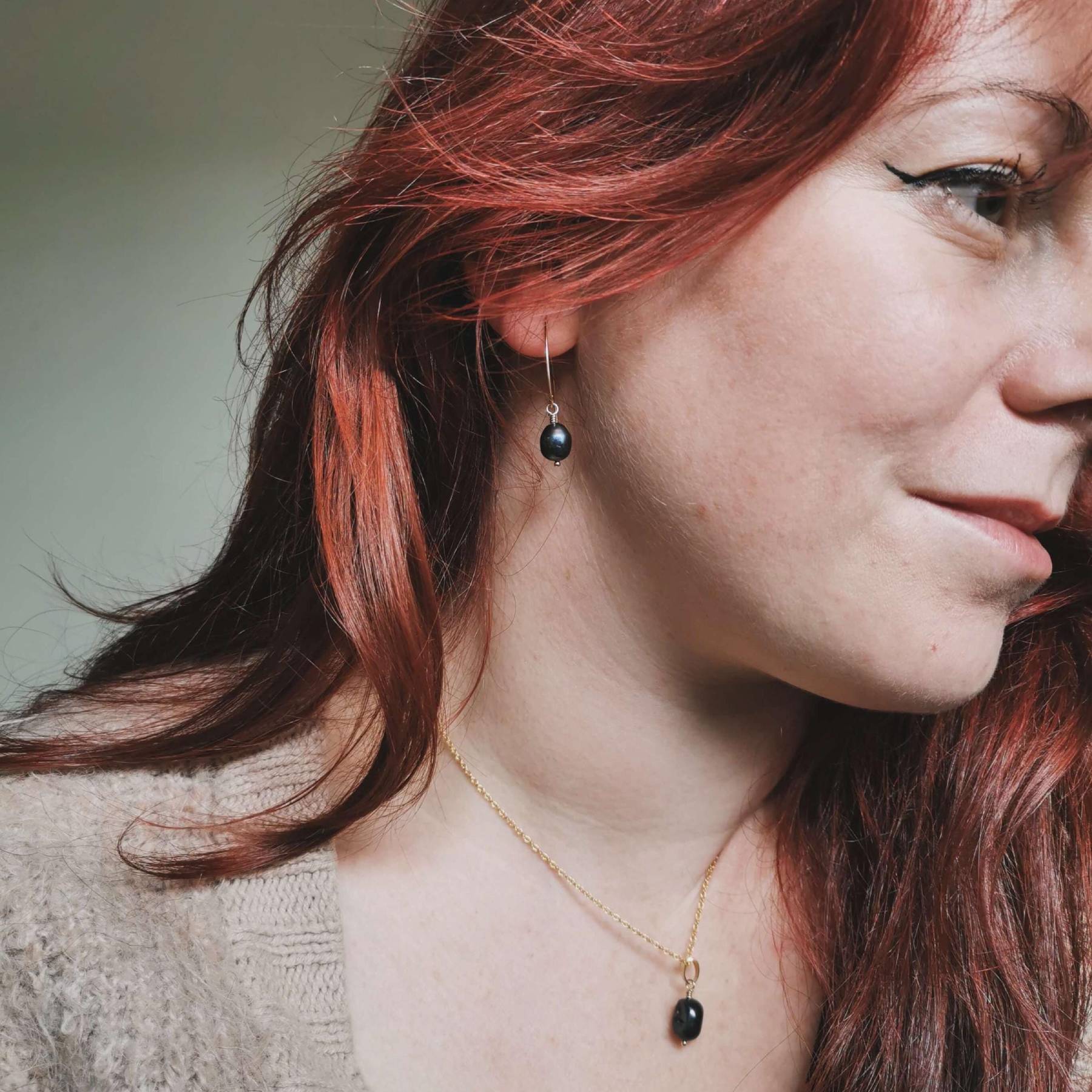 Close-up of a woman with red hair wearing black pearl earrings and a matching gold necklace.
