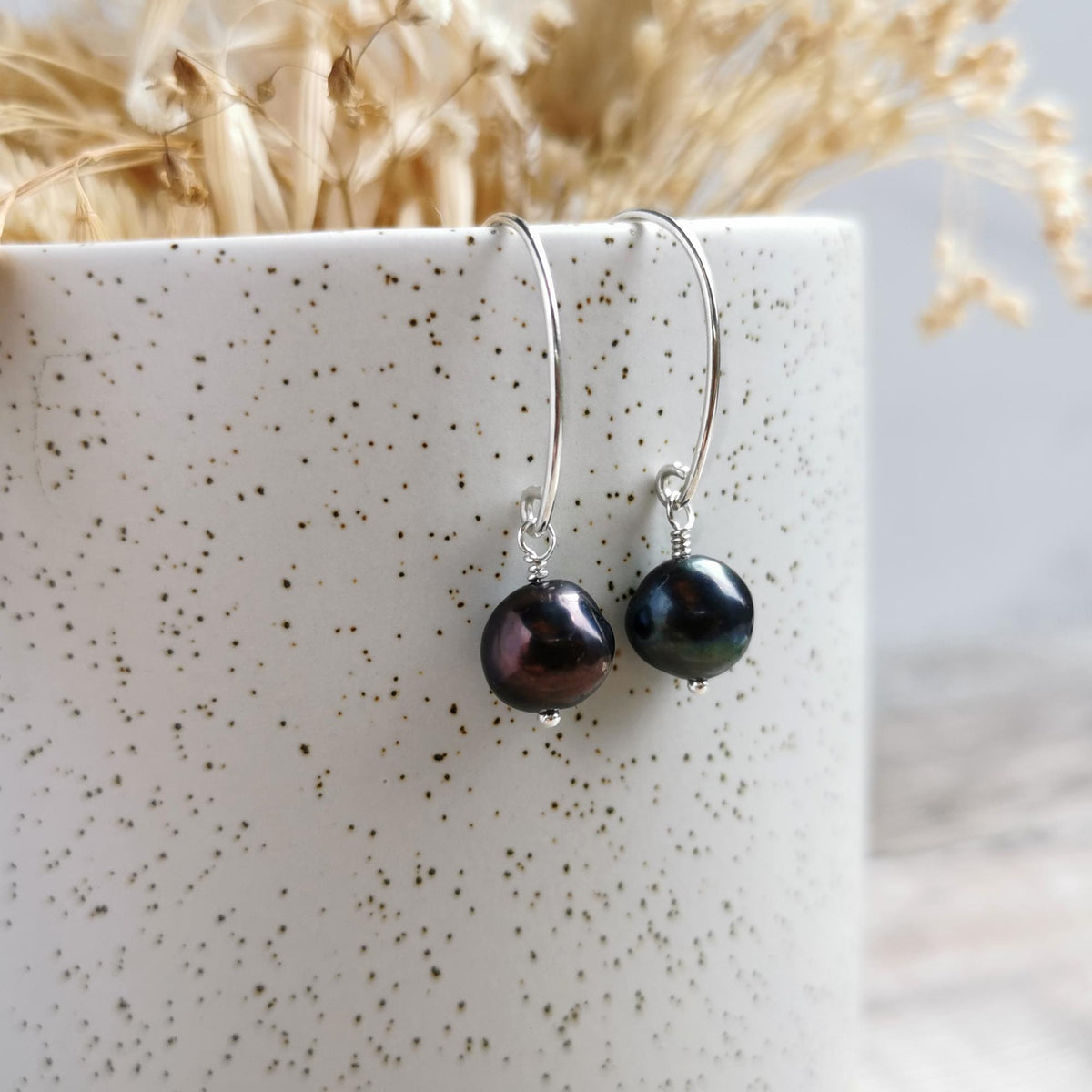 A pair of silver earrings with dark, iridescent peacock purple-black baroque pearl drops, displayed hanging from the rim of a speckled white ceramic cup with dried flowers in the background.