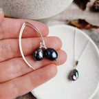 Silver drop earrings with black pearls held in a hand against a neutral background with the matching silver and black pearl necklace