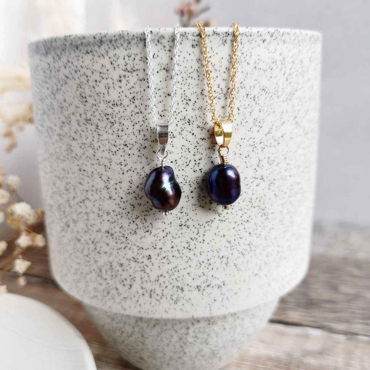 Two handmade peacock baroque pearl necklaces hanging from a speckled ceramic cup. One necklace has a silver-chain and bail with a dark blue-black pearl, and the other has a 14k gold-filled chain and bail with a similar dark pearl.