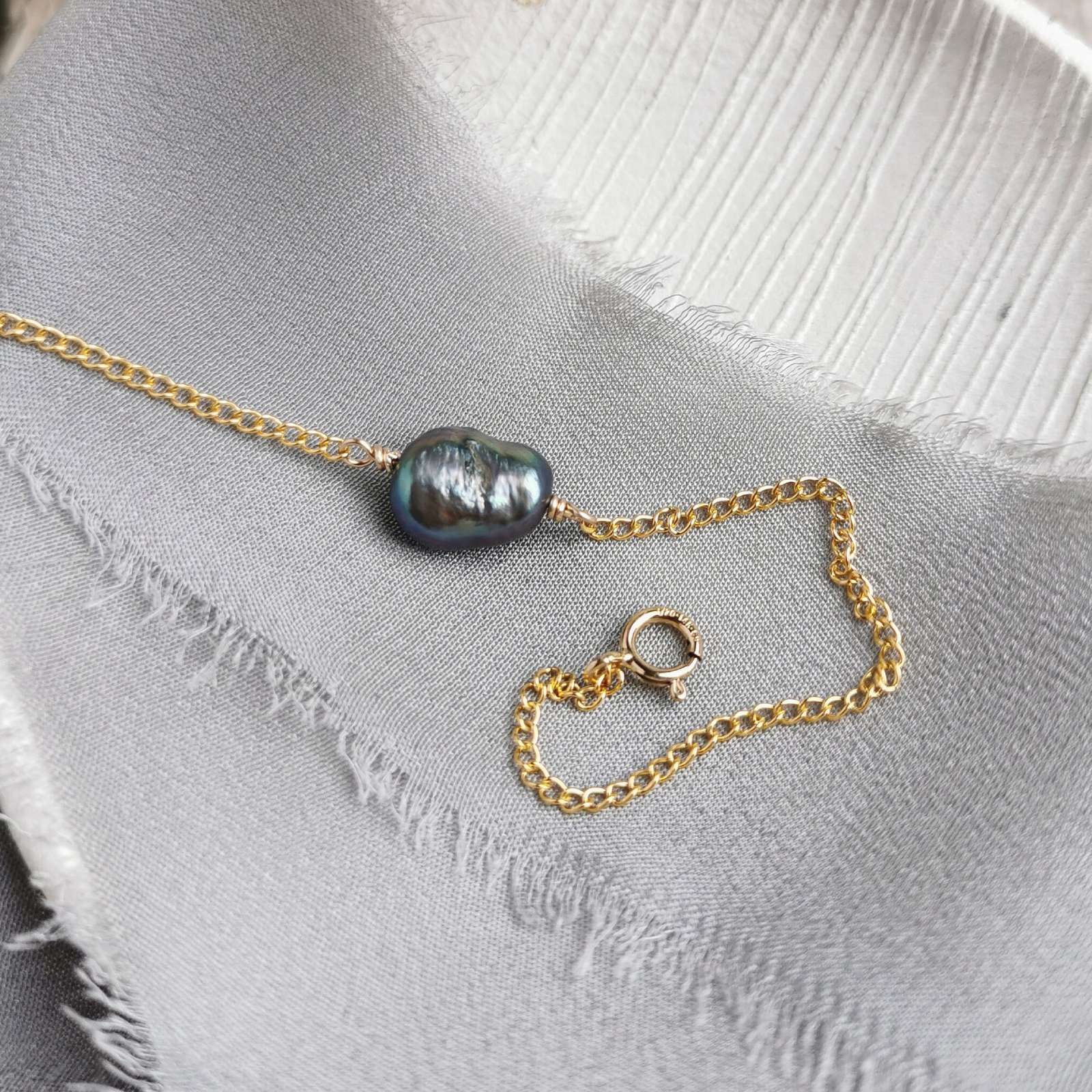 Gold chain bracelet with a large black peacock purple baroque pearl on a white fabric background