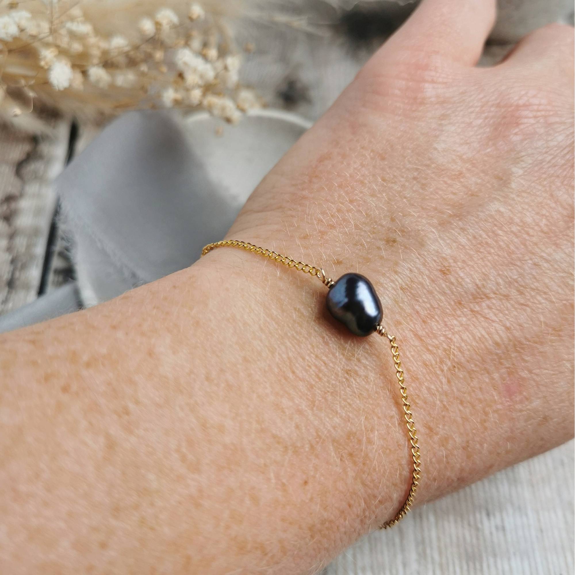 Gold bracelet with a black peacock purple pearl worn on a wrist, blurred background