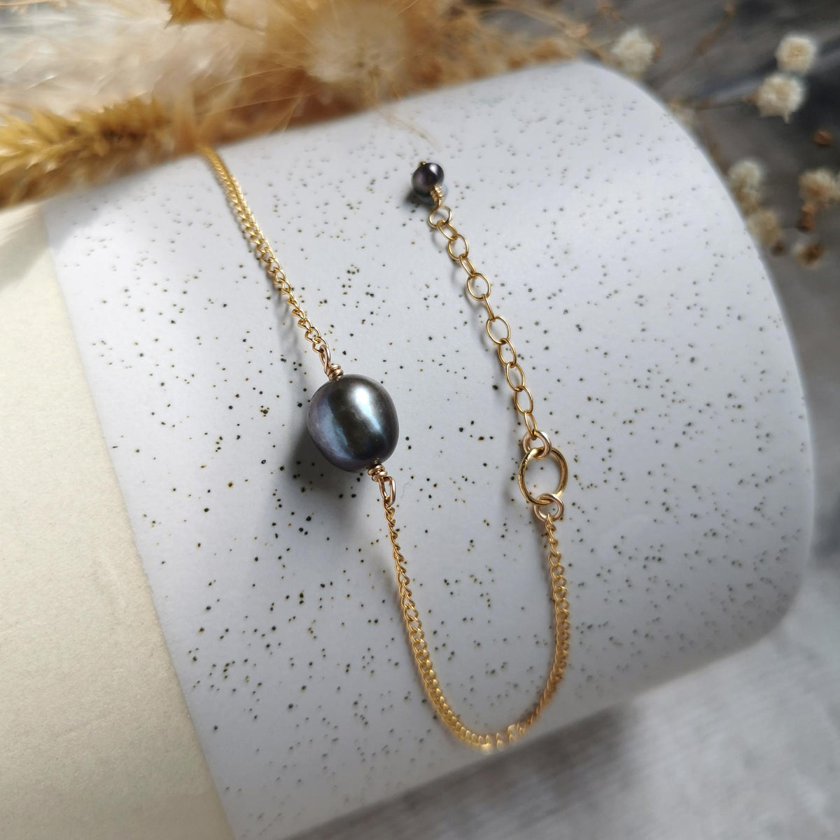 A delicate gold chain bracelet with a single dark purple-gray baroque pearl and an extender chain with smaller matching colour pearl, displayed on a white speckled cylindrical stand.
