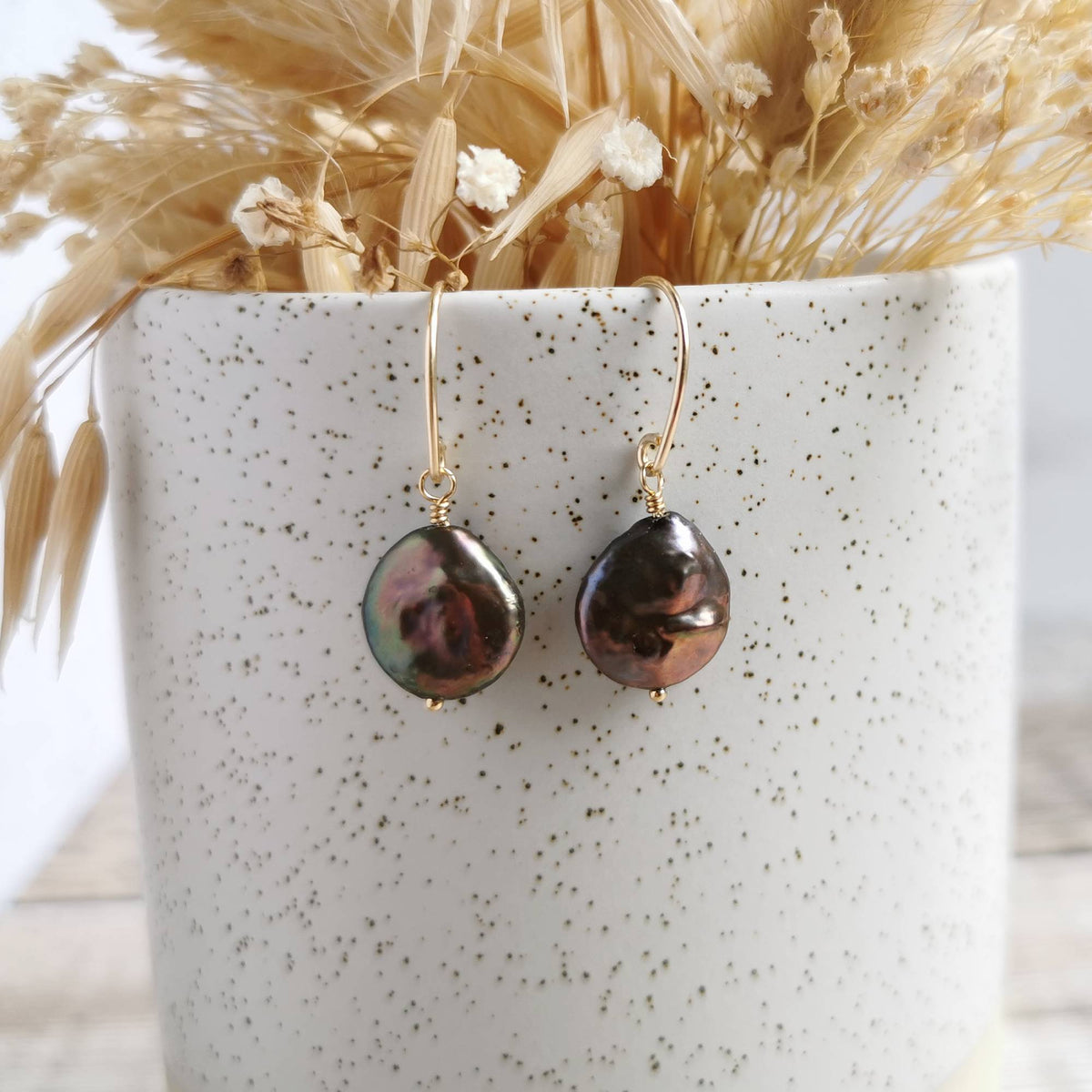 A pair of handmade 14k gold-filled drop earrings with irregular baroque coin pearls in iridescent purple, bronze, and green tones, displayed on a speckled white ceramic pot with dried grass.