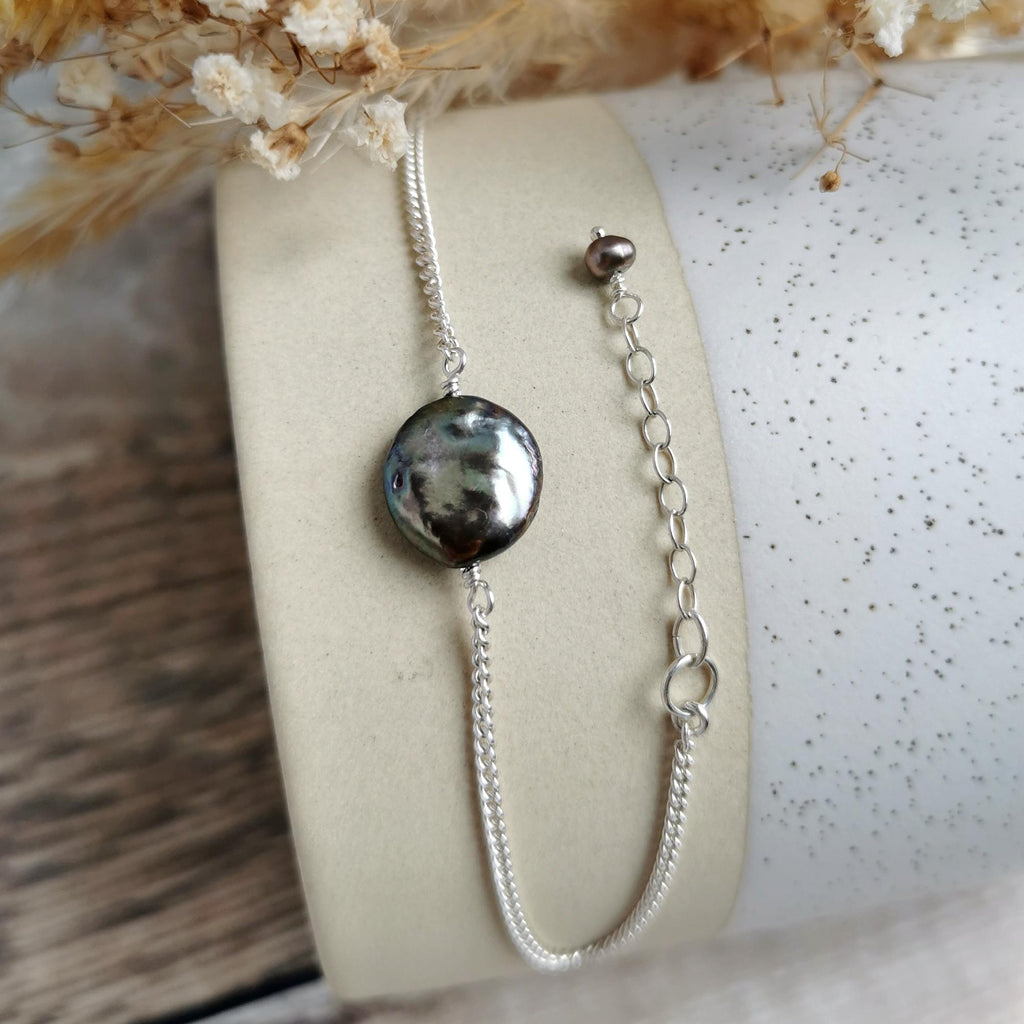 A silver chain bracelet with an irregular dark gray and black coin pearl and matching extender chain, displayed on a cream-colored cylindrical stand.
