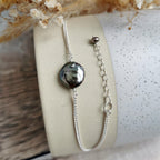 A silver chain bracelet with an irregular dark gray and black coin pearl and matching extender chain, displayed on a cream-colored cylindrical stand.