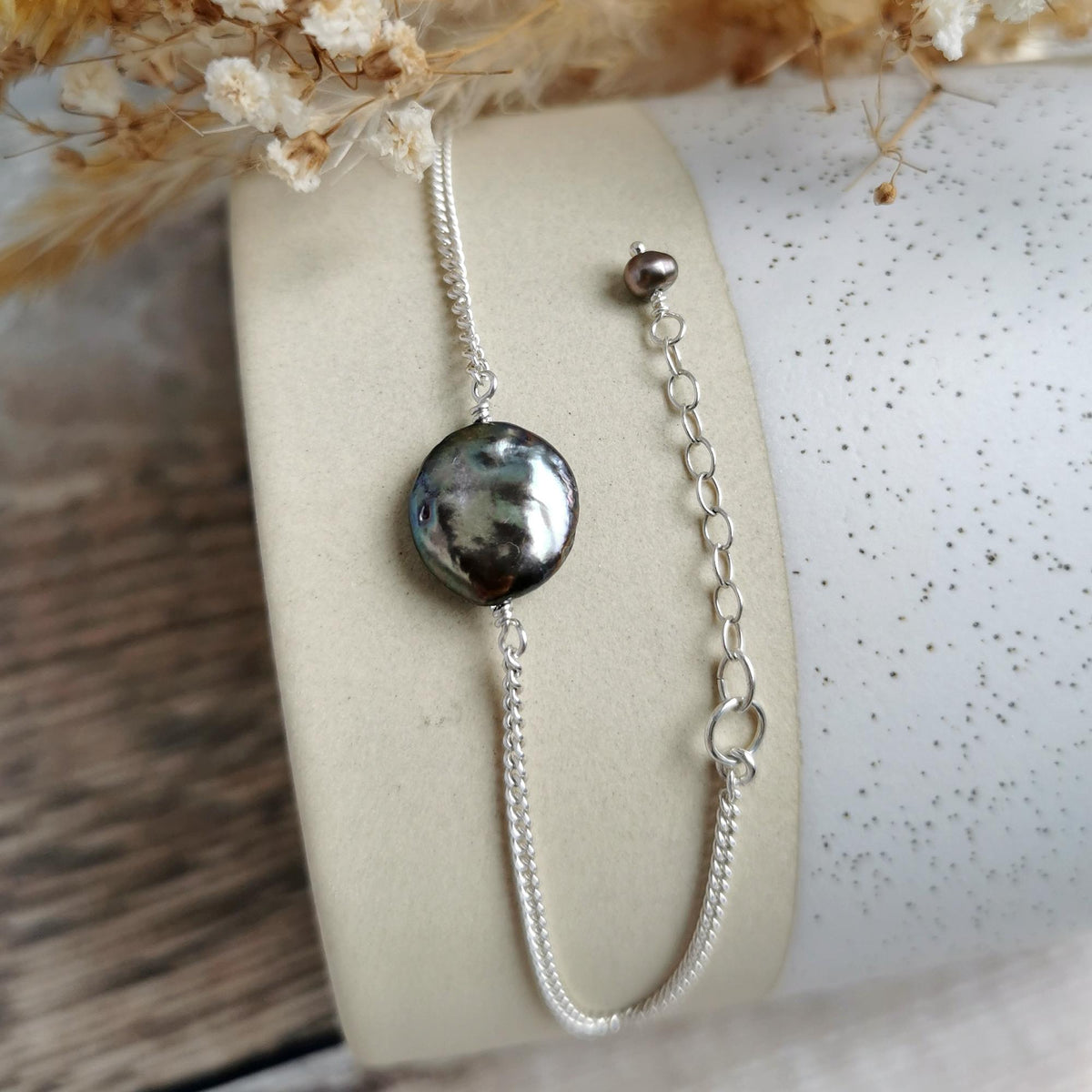 A silver chain bracelet with an irregular dark gray and black coin pearl and matching extender chain, displayed on a cream-colored cylindrical stand.