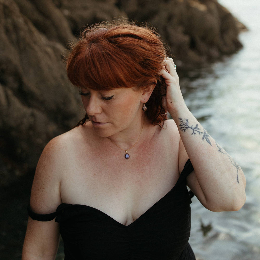 A woman with red hair wearing a black swimsuit and a minimal gold and peacock coin pearl necklace, standing outdoors near the sea and rocks, looking down and holding her hair back to reveal the matching peacock pearl earrings.