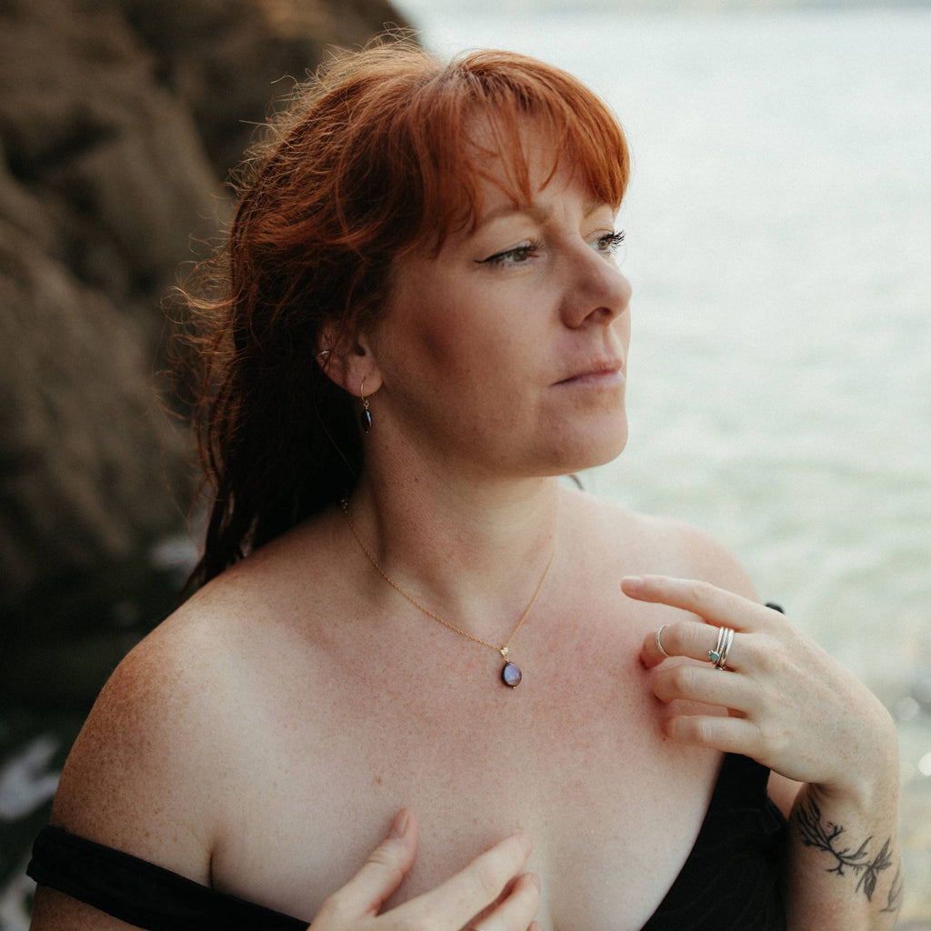A woman with red hair wearing a delicate gold necklace with a small peacock purple pearl pendant, posing outdoors near the water with a blurred background of rocks and sea in cornwall.