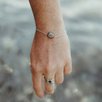 A close-up of a person's hand and wrist wearing a delicate silver chain bracelet with a round peacock pearl, set against a blurred outdoor background over the sea in cornwall