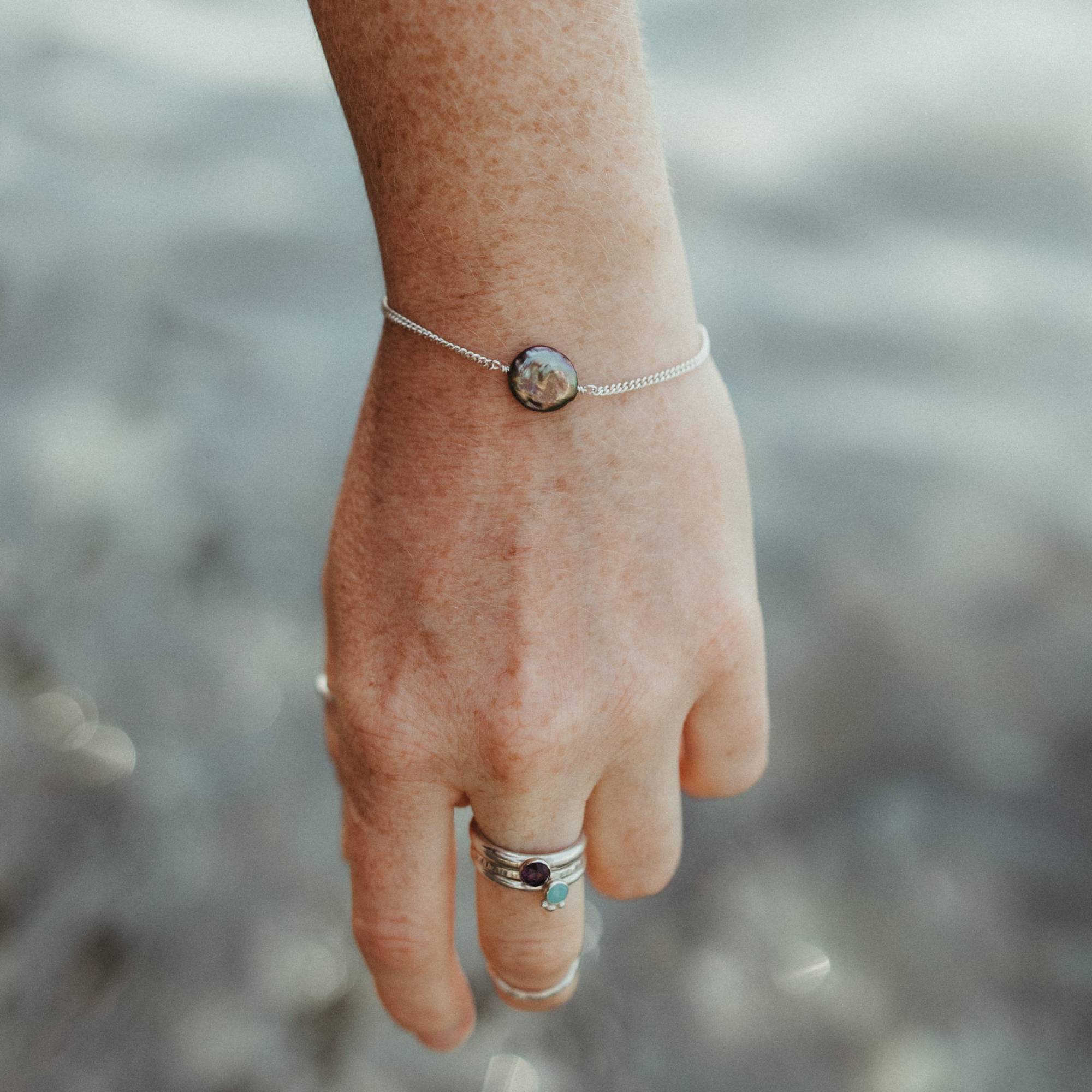 A close-up of a person's hand and wrist wearing a delicate silver chain bracelet with a round peacock pearl, set against a blurred outdoor background over the sea in cornwall
