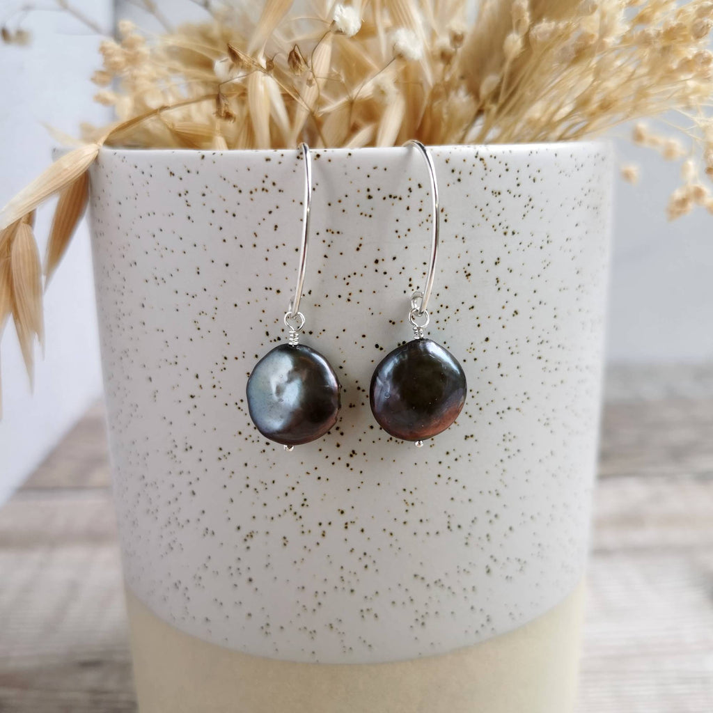 A pair of handmade silver earrings with round, dark baroque coin pearls, displayed on a speckled ceramic cup filled with dried grass.