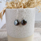 A pair of handmade silver earrings with round, dark baroque coin pearls, displayed on a speckled ceramic cup filled with dried grass.