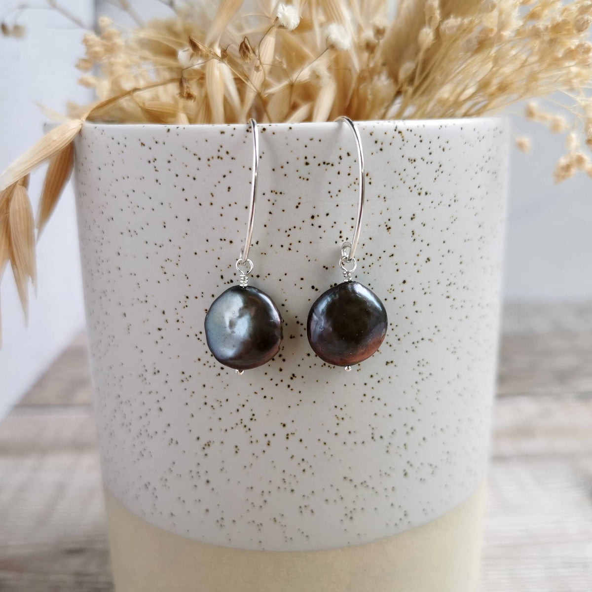 A pair of handmade silver earrings with round, dark baroque coin pearls, displayed on a speckled ceramic cup filled with dried grass.