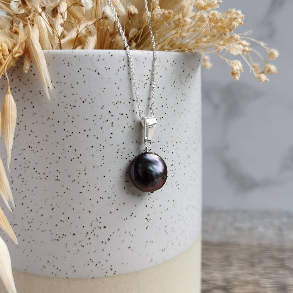 A silver pendant necklace featuring a lustrous dark pearl, draped over a speckled white ceramic vase with dried flowers.