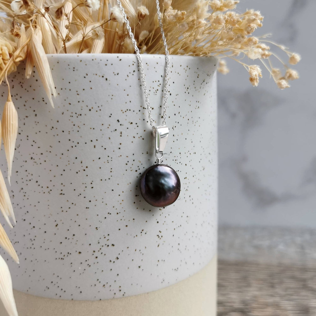 A silver pendant necklace featuring a lustrous dark pearl, draped over a speckled white ceramic vase with dried flowers.