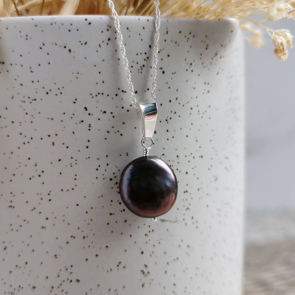 A silver pendant necklace featuring a single round, dark purplish blue coin shaped freshwater pearl, draped over a speckled white ceramic vase.