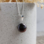 A silver pendant necklace featuring a single round, dark purplish blue coin shaped freshwater pearl, draped over a speckled white ceramic vase.