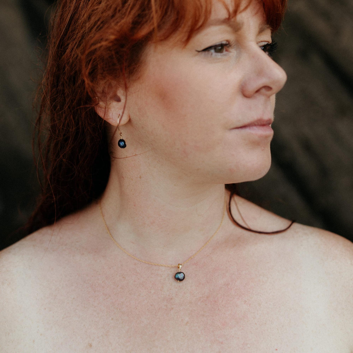 A close-up side profile of a woman with red hair wearing a delicate 14k gold-filled necklace and matching drop earrings with dark blue-purple baroque pearls with an iridescent shimmer.