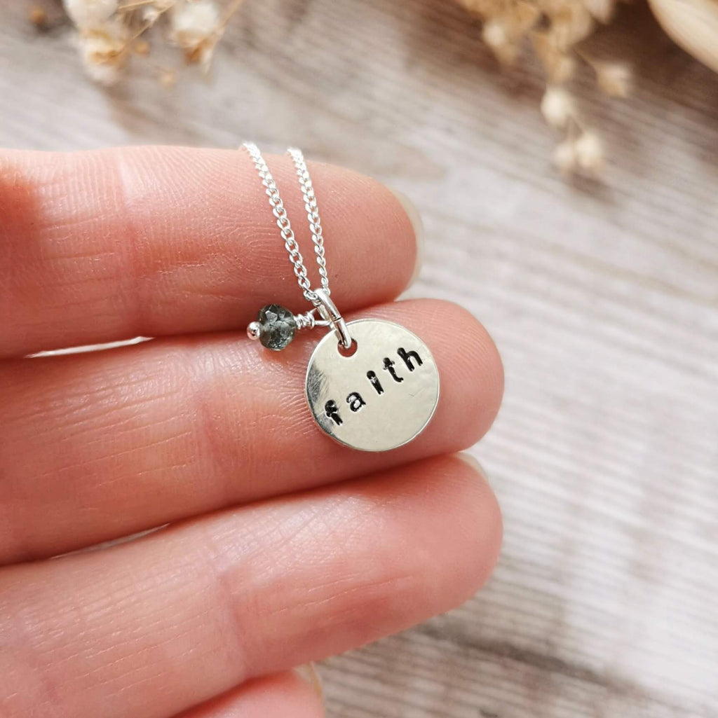 A silver necklace with a round disc pendant hand-stamped with the word 'faith' and a small dark teal moss aquamarine bead charm, held on a person's fingers against a blurred wooden background.