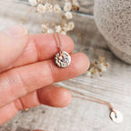A close-up of a hand holding a delicate necklace with a textured round rose gold pendant and small pearl and tanzanite birthstone charms on a fine chain, set against a rustic wooden background with dried flowers.