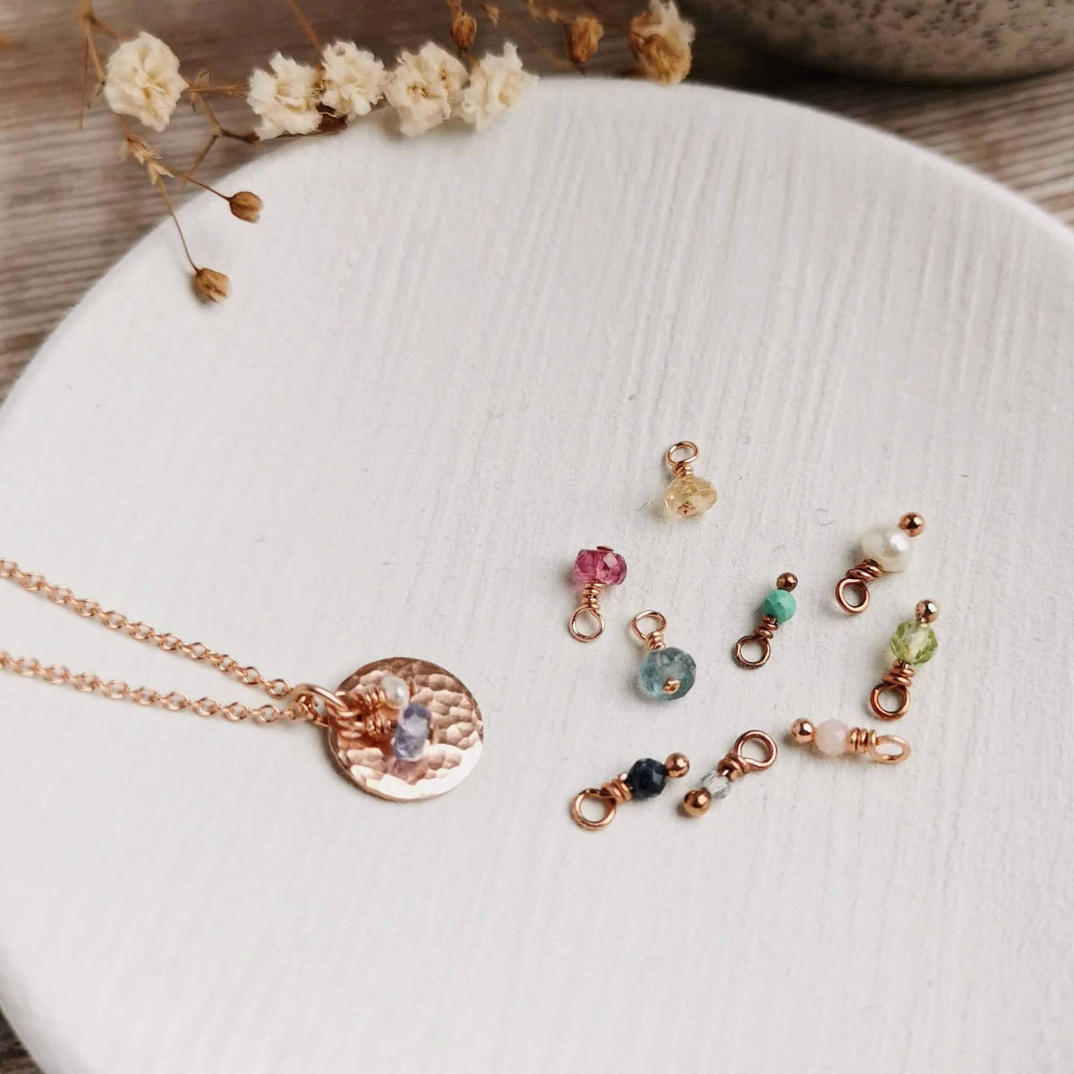 hammered rose gold pendant on a white ceramic dish with gemstone charms attached and scattered around for all birth months, with neutral background and dried flowers