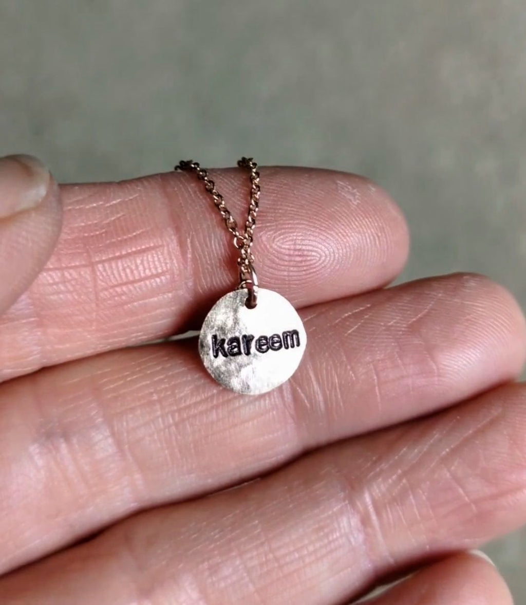 A close-up of a hand holding a small round rose gold pendant necklace stamped on the back with the name 'kareem', hung on a delicate rose gold chain.