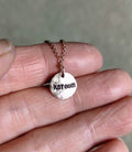 A close-up of a hand holding a small round rose gold pendant necklace stamped on the back with the name 'kareem', hung on a delicate rose gold chain.