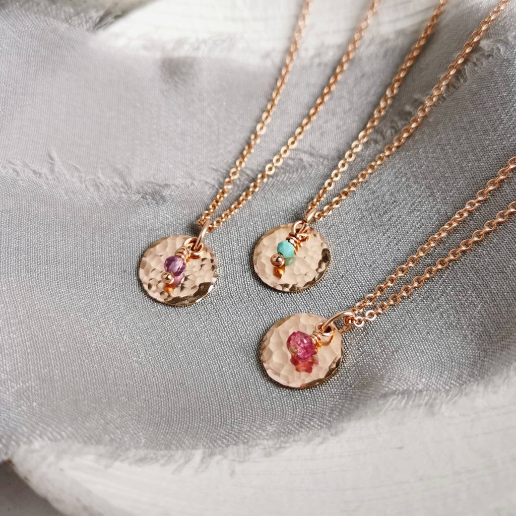Three rose gold birthstone necklaces with hammered round pendants and small colorful gemstones for february (amethyst), Turquoise (december) and pink tourmaline (october), arranged on a textured light gray fabric background.