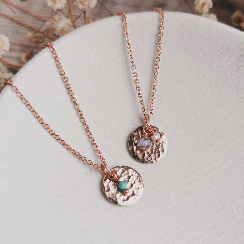 Two rose gold birthstone necklaces with hammered round pendants, one featuring a small turquoise bead for december and the other a small tanzanite bead as an alternative gemstone for december with a small pearl for june, displayed on a textured white surface.