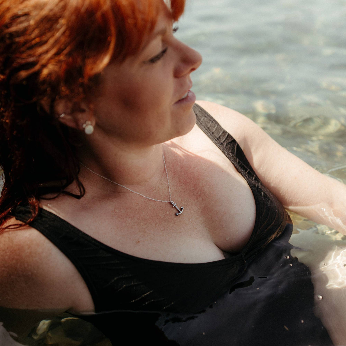 A woman with red hair wearing a black swimsuit and a silver anchor necklace, sitting in shallow water.