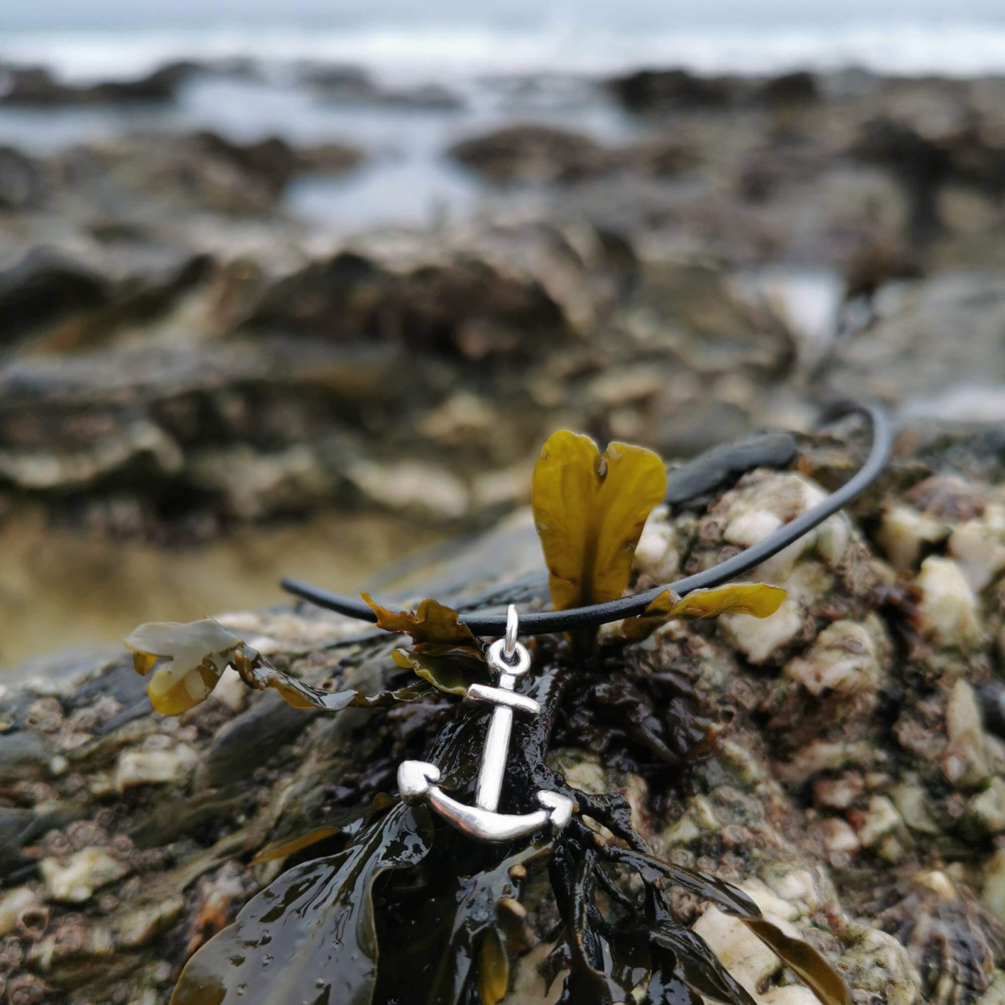 Necklace with a silver anchor charm and black leather cord, laid on seaweed by the sea in cornwall