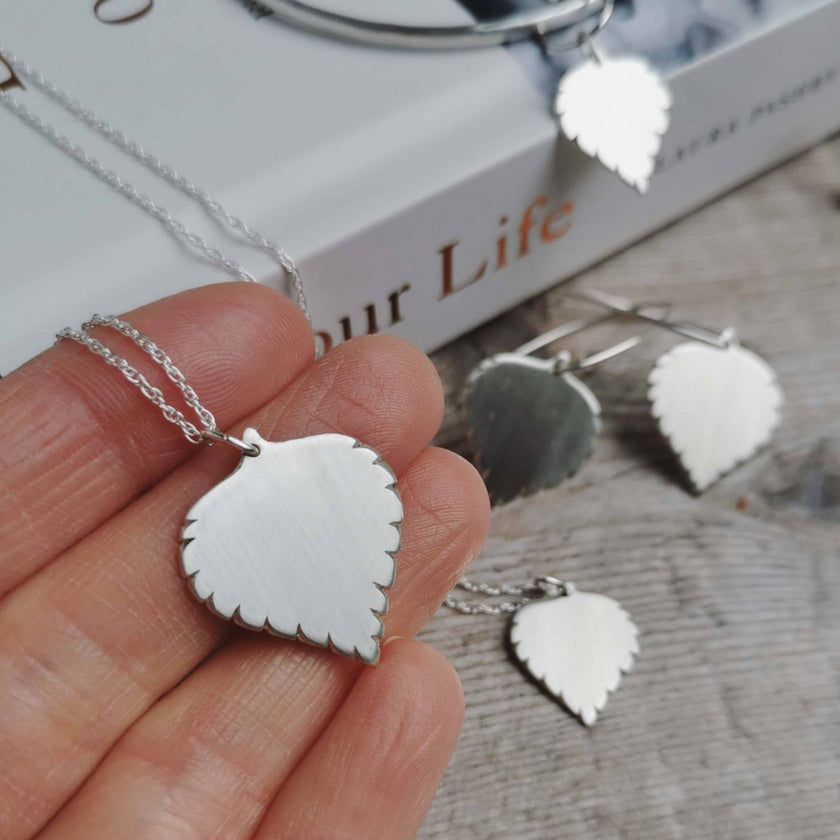 large handmade silver birch leaf necklace held in hand with the rest of the jewellery collection blurred in the background