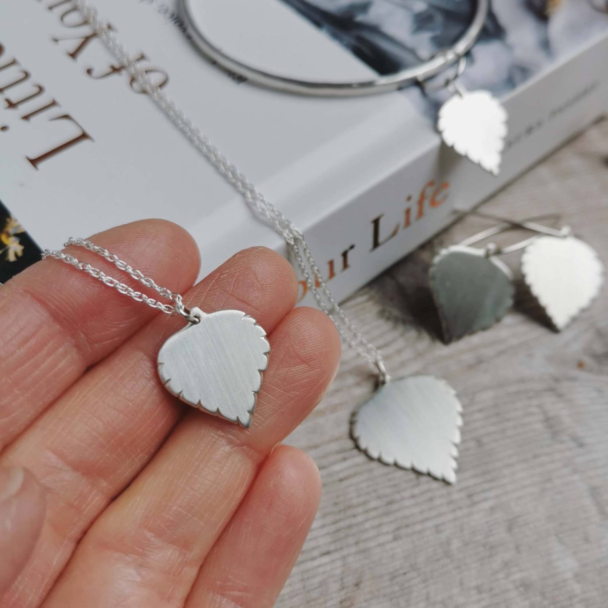 small handmade silver birch leaf necklace held in hand with the rest of the silver jewellery collection blurred in the background