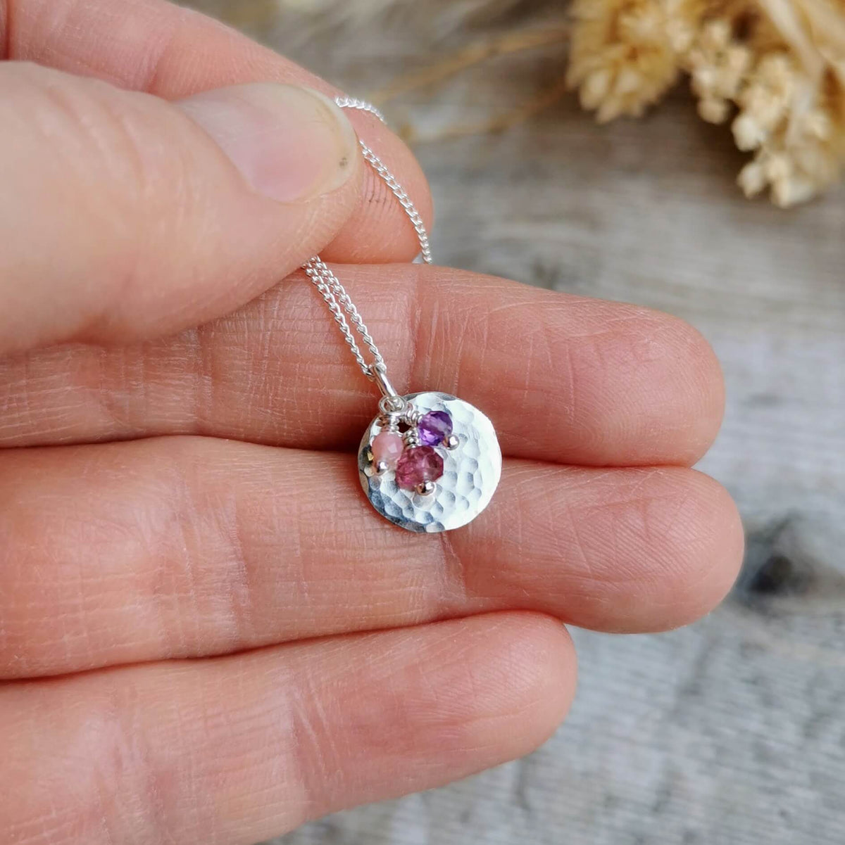 A close-up of a hand holding a silver pendant birthstone necklace with a hammered disc charm and small purple and pink opal, tourmaline and amethyst gemstones for november, november alternative and february birth months, against a blurred rustic background.