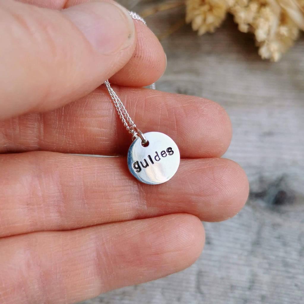 A close-up of a hand holding a small silver round birthstone pendant necklace hand stamped with the word 'guides' hidden on the back.