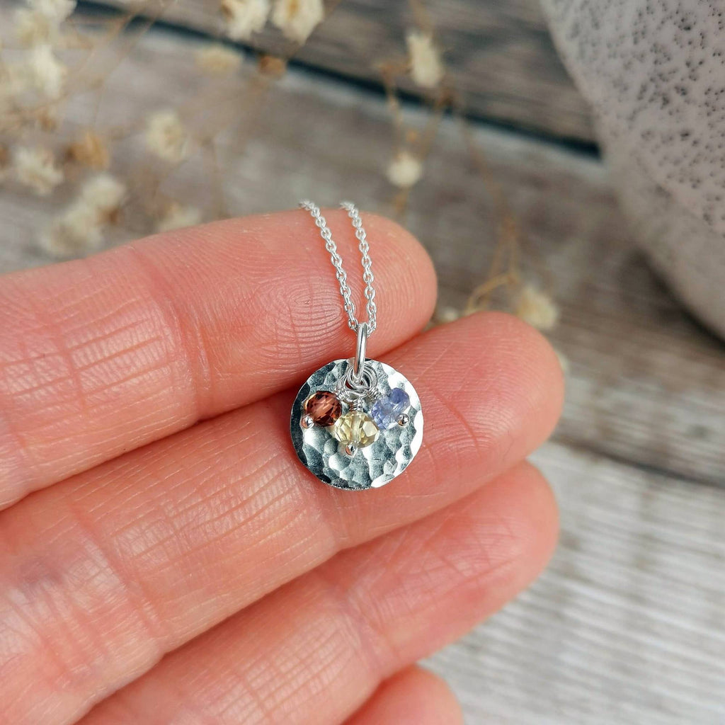 A close-up of a hand holding a small hammered silver pendant birthstone necklace with three tiny colorful gemstones on a silver chain - garnet for january, citrine for october and tanzanite as an alternative gemstone for december, set against a blurred rustic background.