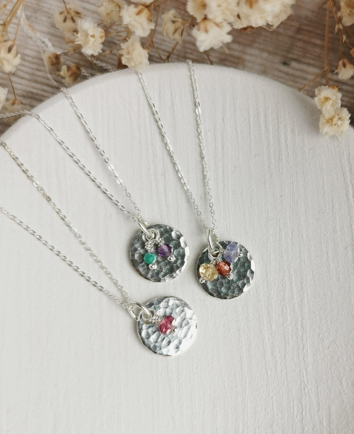 A collection of hammered silver birthstone necklaces with various colored gemstones for February, October, November, and december birth months, displayed on a white surface with dried flowers. 