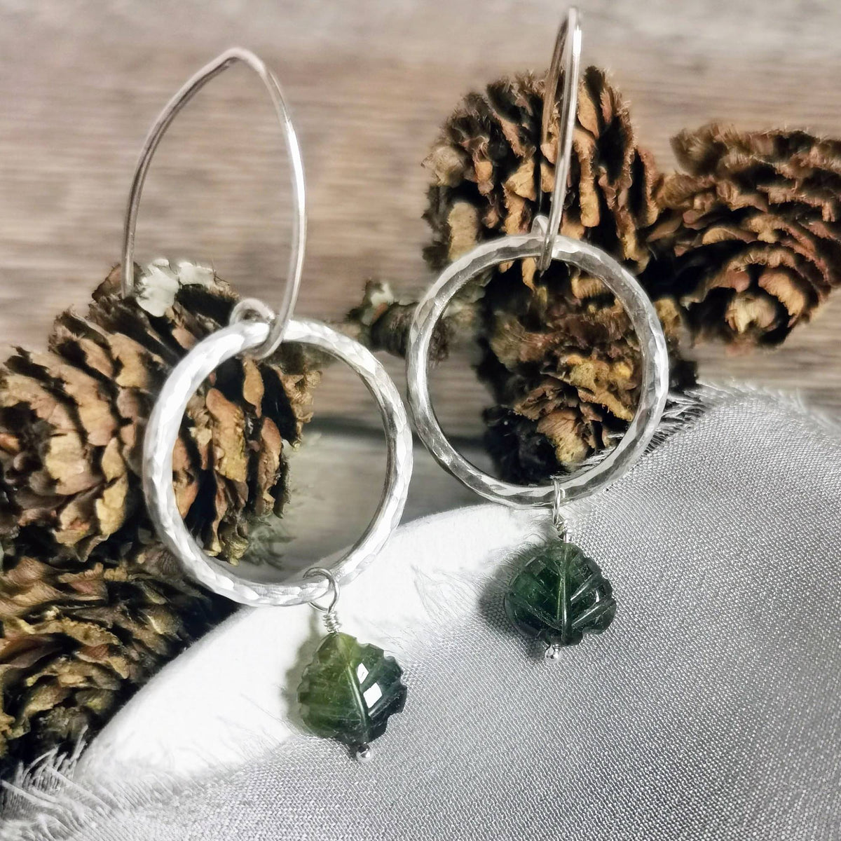 handmade Textured Silver circle earrings with green gemstone leaves on a textured surface styled with pine cones.