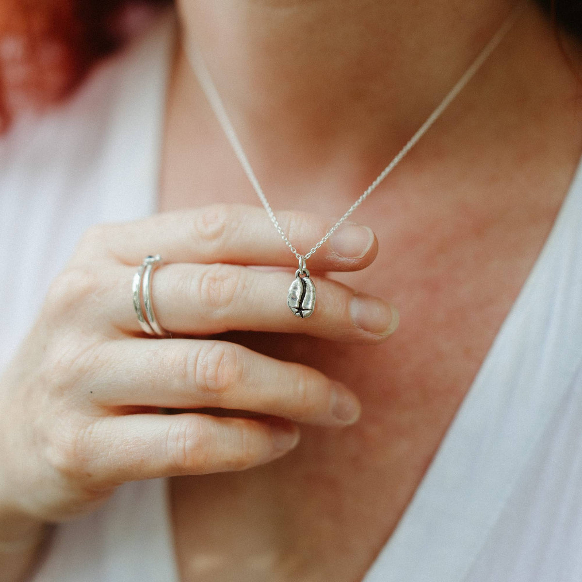 A close-up lifestyle shot of a person wearing a delicate silver coffee bean pendant necklace and silver rings, holding their hand behind the necklace.