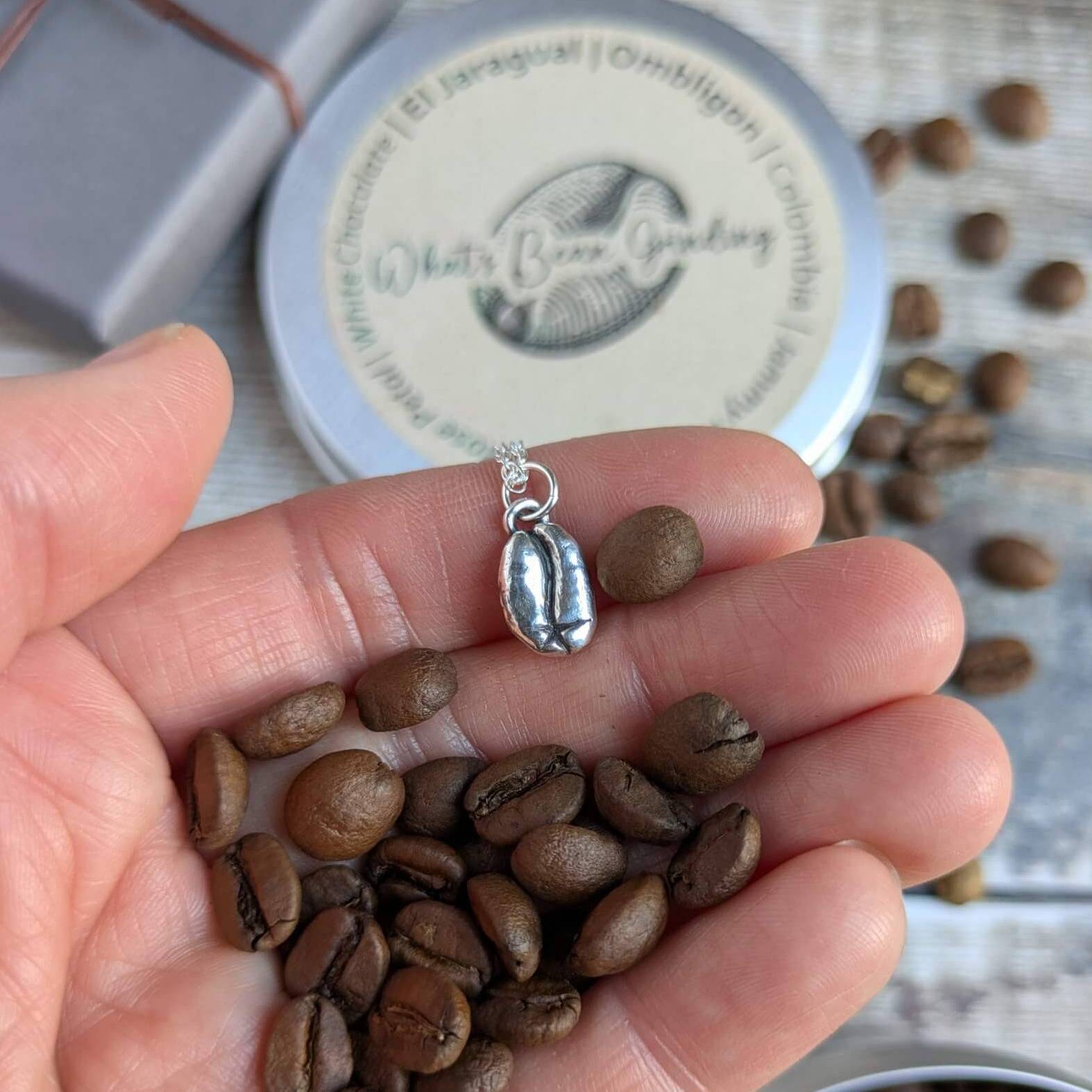 Hand holding coffee beans with a silver coffee bean-shaped charm, and a branded container in the background for the coffee roaster Chris Whitworth at What's Bean Grinding