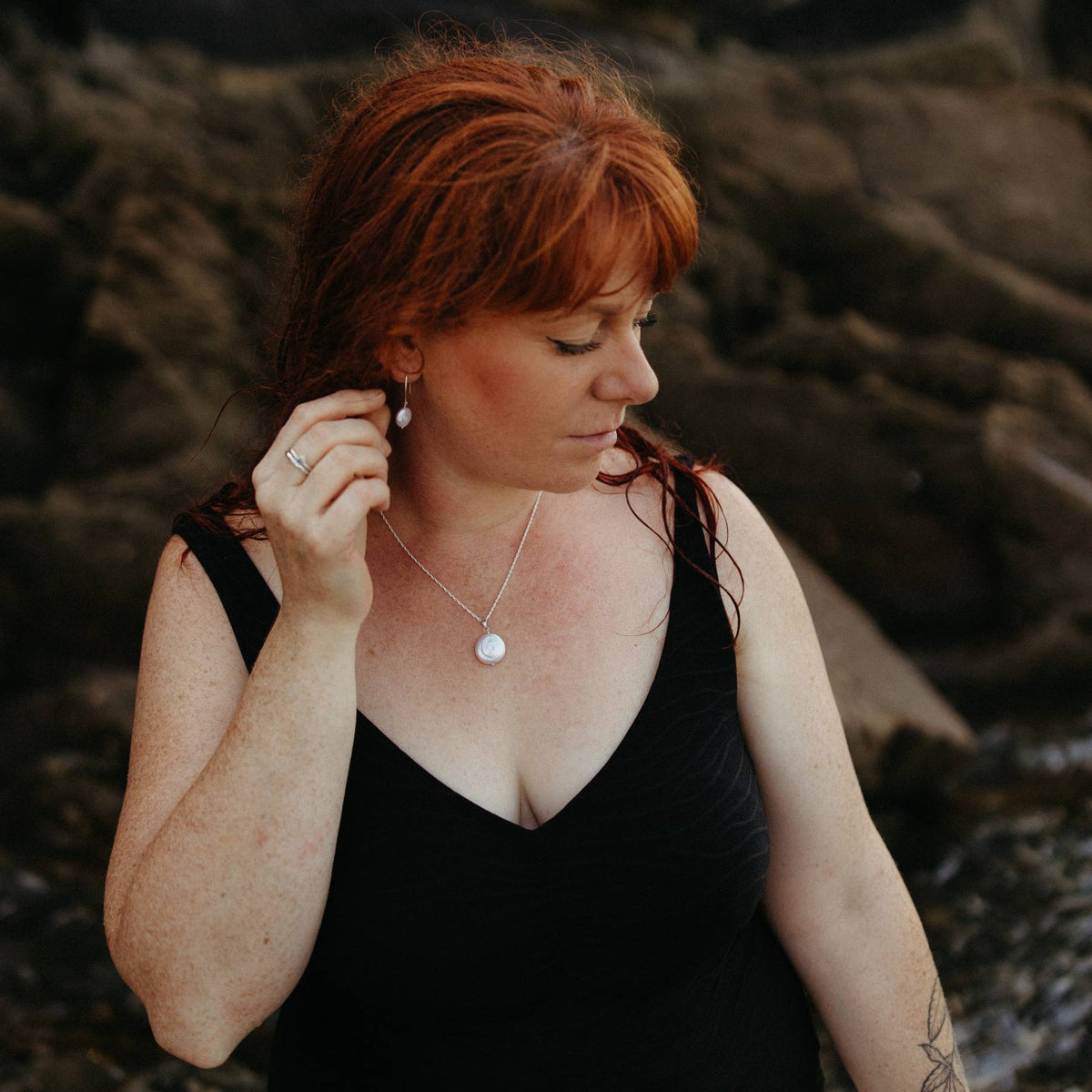 A woman with red hair wearing a black swimsuit and a silver and pearl statement necklace, and matching smaller coin pearl earrings, standing outdoors against a blurred rocky coastal background in Cornwall, UK.