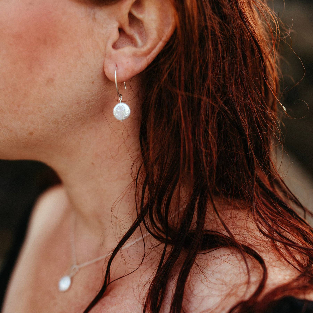 A close-up lifestyle shot of a woman wearing delicate silver ivory coin pearl earrings and matching necklace. The warm, reddish-brown hair frames the side of her face and ear, with a soft-focus dark background.