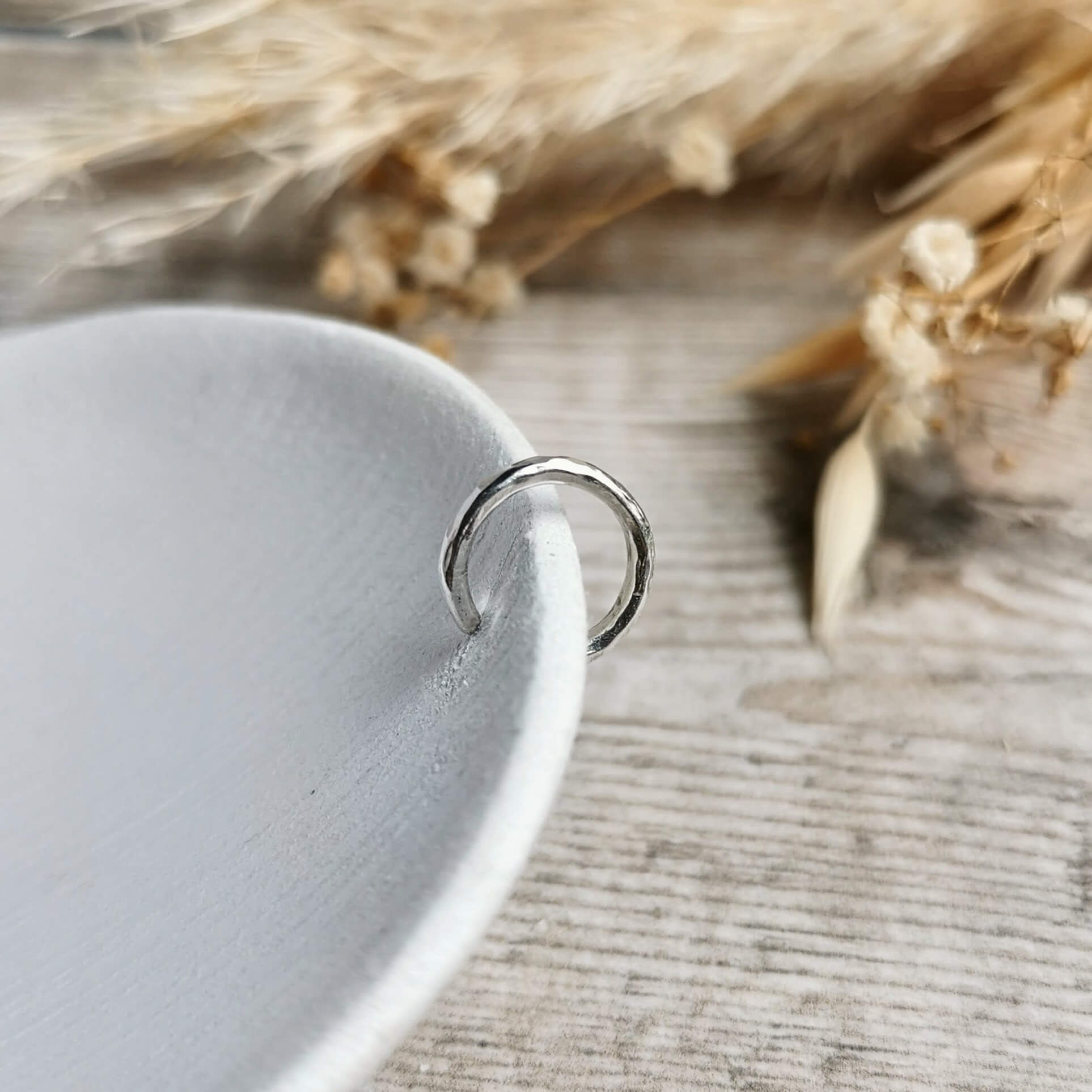 Close-up of a hammered silver ear cuff on a textured surface with dried plants in the background