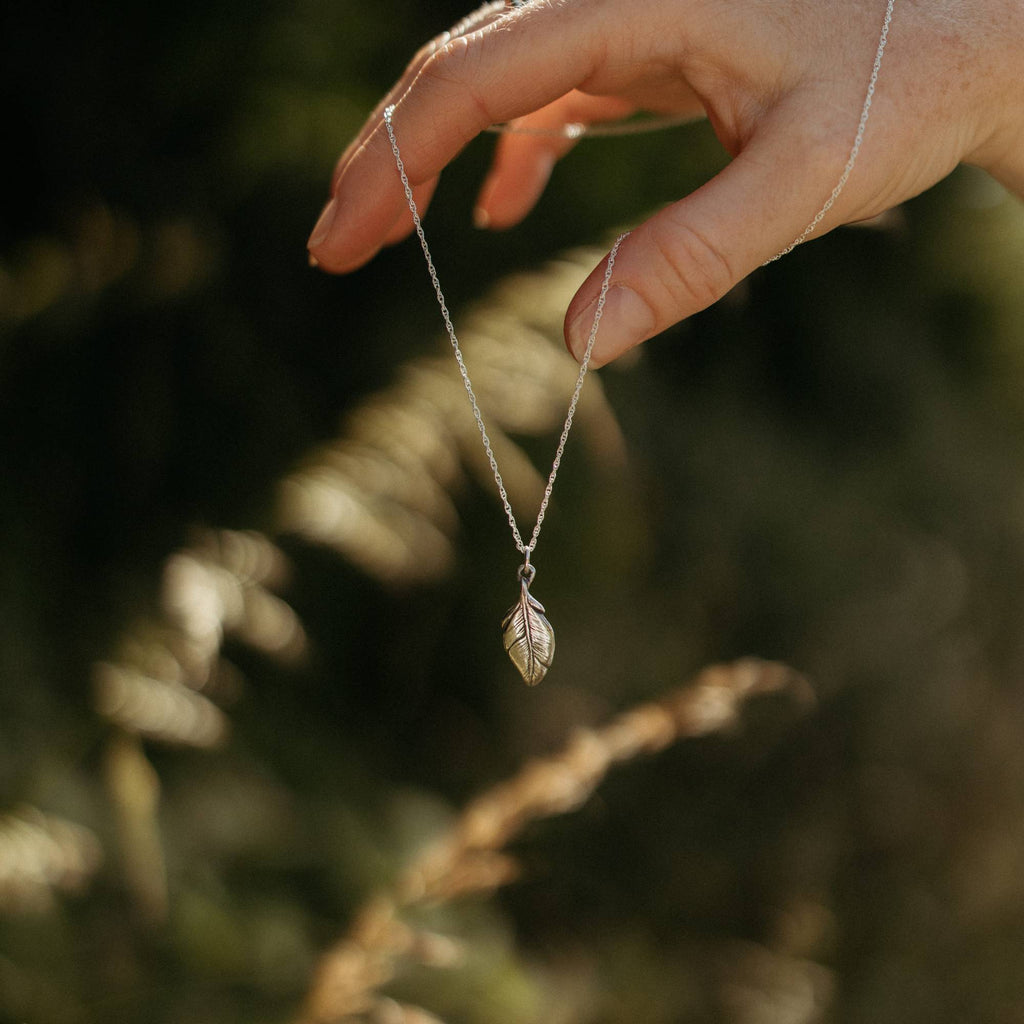 A delicate silver necklace with a small feather pendant is held by a hand against a blurred outdoor background of green foliage and warm sunlight.